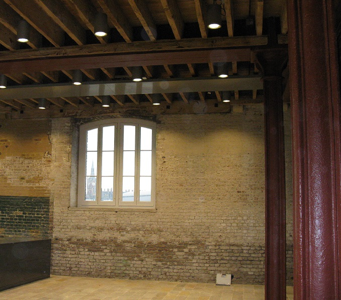 View inside a vacant workshop showing bare brick wall, arched window, and cast iron pillars supporting ceiling timbers